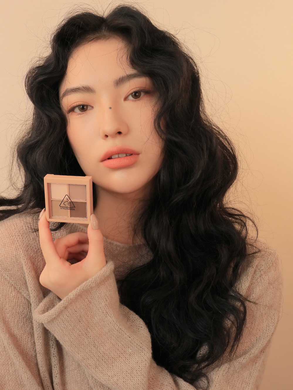 Model wearing a cozy beige sweater holding 3CE Mini Multi Eye Color Palette in Shade Flat Call, demonstrating its application and wearable look.