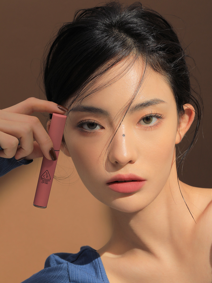 Model wearing a blue top against an earthy brown background featuring 3CE Velvet Lip Tint in Shade Cashmere Nude, emphasizing its soft texture and natural matte look.