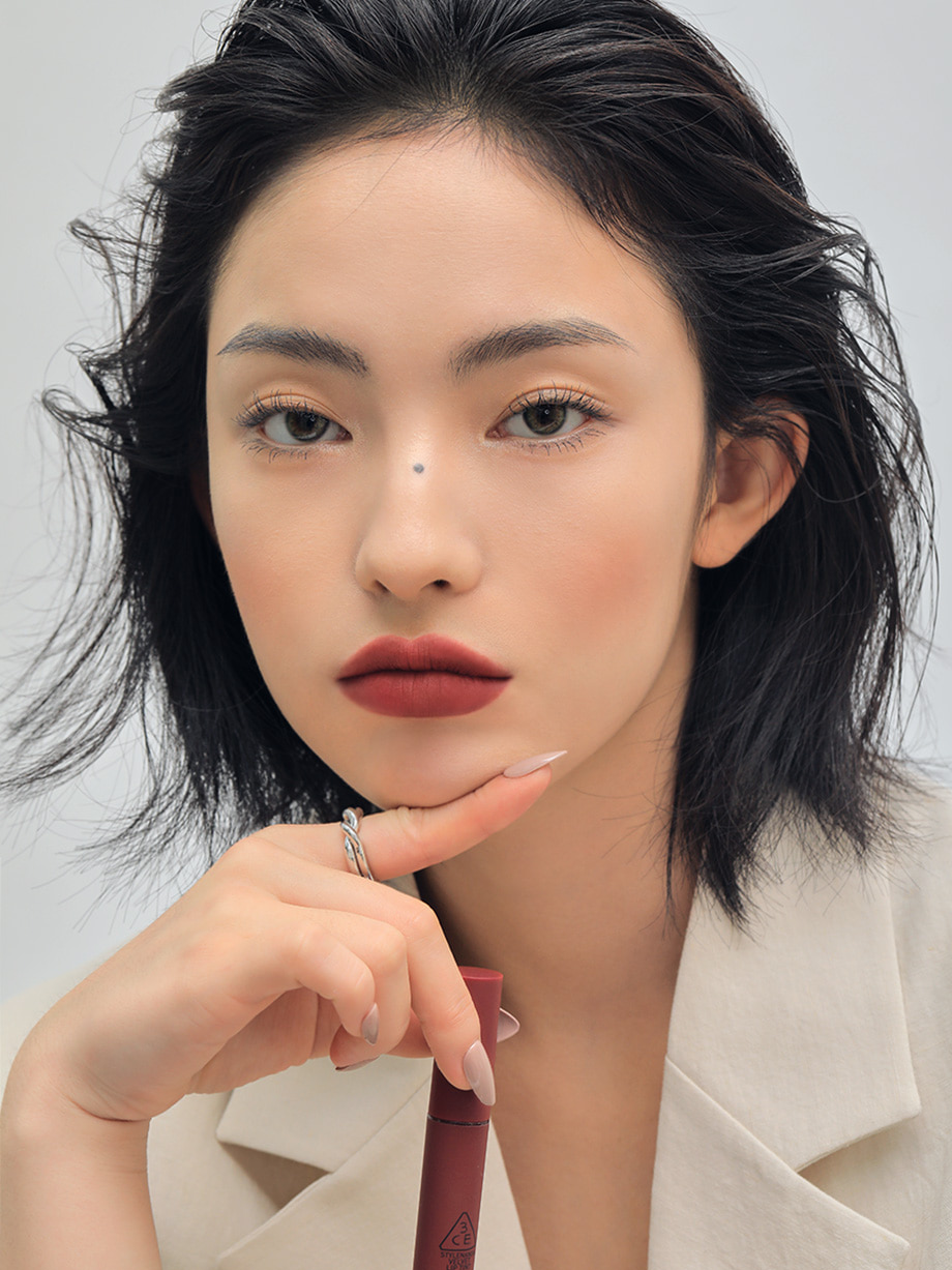 Model with shoulder-length wavy hair wearing 3CE Velvet Lip Tint in Shade Definition, showcasing bold pigmentation and a velvety matte finish.