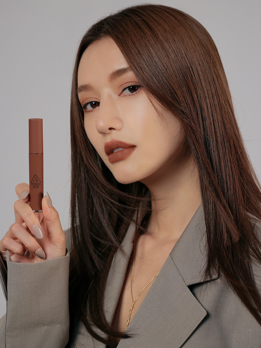 Model with long, sleek brown hair in a stylish gray blazer applying 3CE Velvet Lip Tint in Shade Bitten Hour, highlighting its velvety finish and intense color payoff.