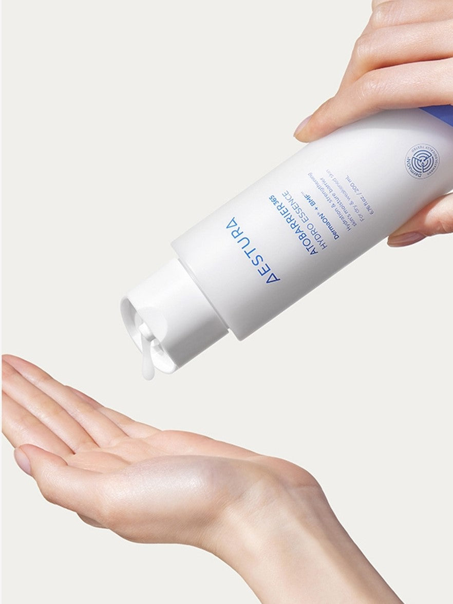 A hand dispensing milky-light hydrating essence from an Aestura Atobarrier 365 Hydro Essence bottle onto another hand, set against a light background.