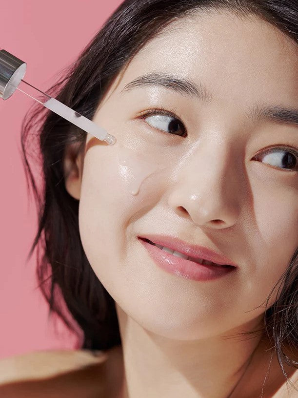 A model holds an Aestura Theracne 365 Active Serum dropper above her skin, with a soft pink background emphasizing the product’s light texture.