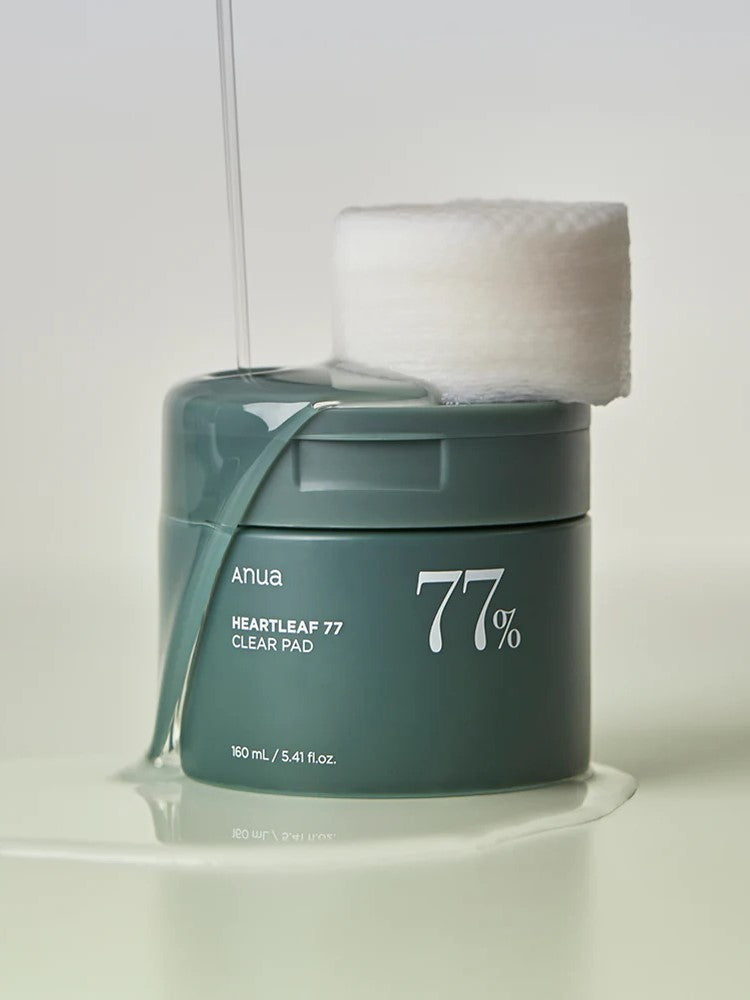 A sleek green jar labeled 'Anua Heartleaf 77% Clear Pad' with cotton pads resting on top, surrounded by a glossy liquid spill that highlights the essence of the pads.