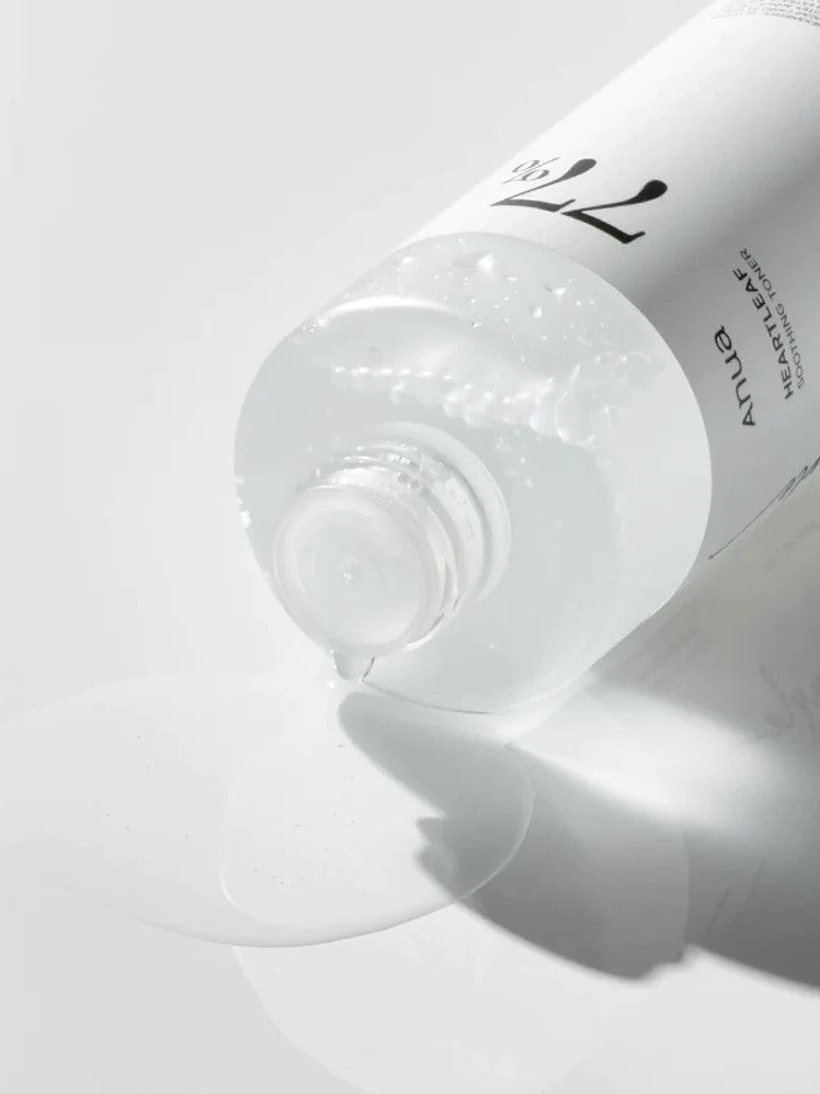 A partially tipped bottle of Anua Heartleaf 77% Soothing Toner spills a few drops onto a white surface, showcasing its lightweight texture.