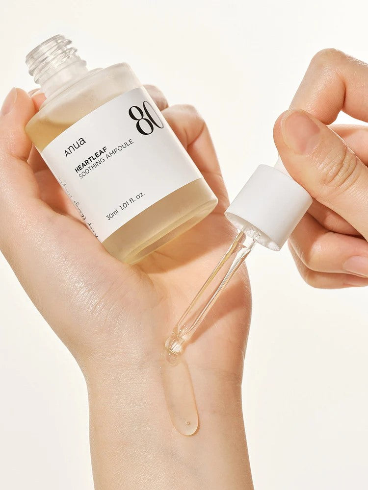 A hand holds a bottle of Anua Heartleaf Soothing Ampoule, with a dropper dispensing liquid onto the wrist.