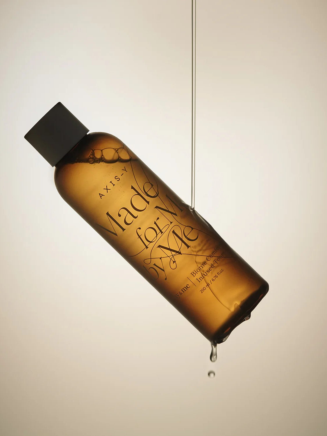 AXIS-Y Biome Comforting Infused Toner, suspended in air with a droplet falling from the bottom against a soft beige backdrop.
