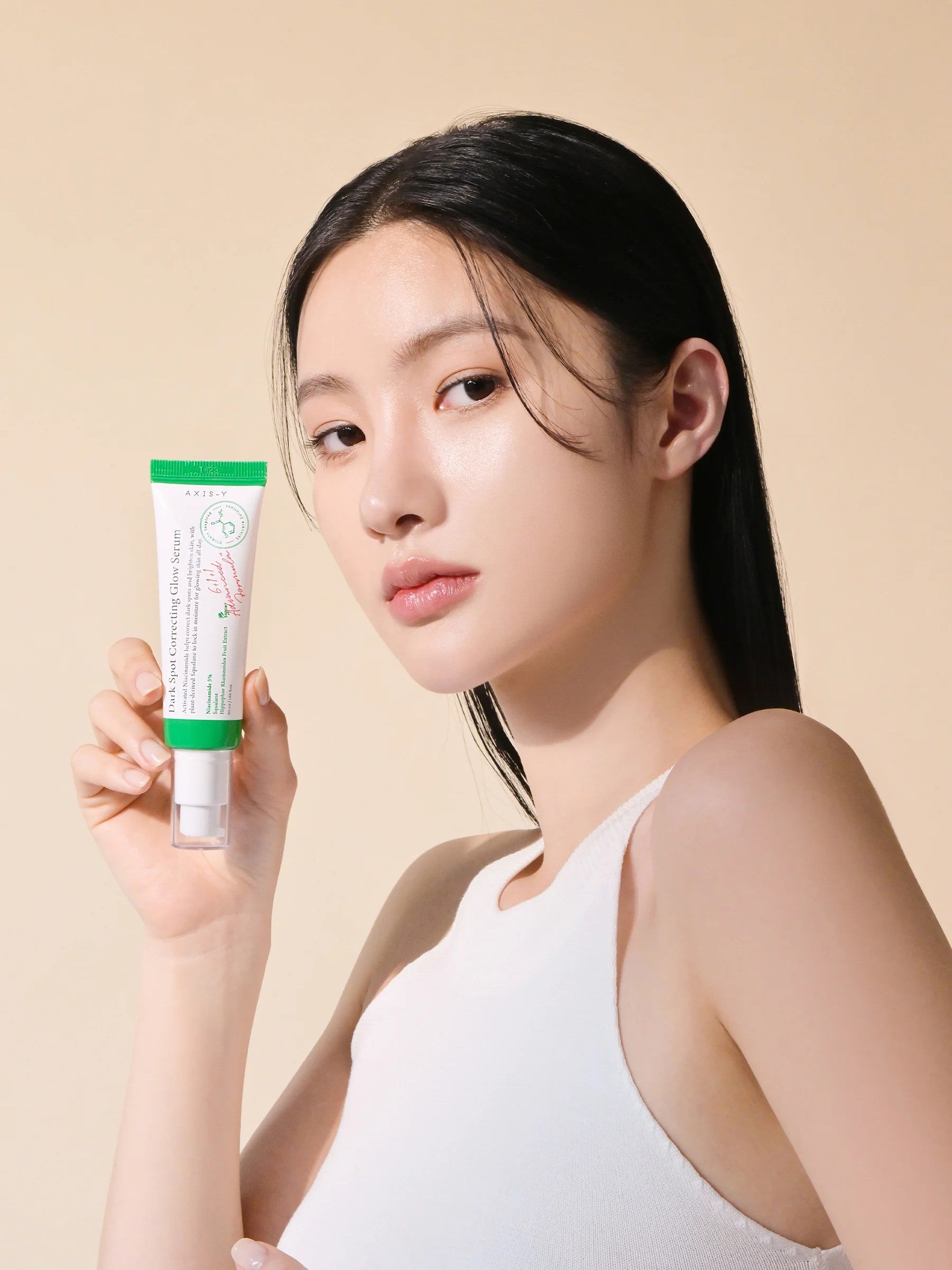 A model with long black hair holds Axis-Y Dark Spot Correcting Glow Serum against a light beige background, showcasing the product’s design.