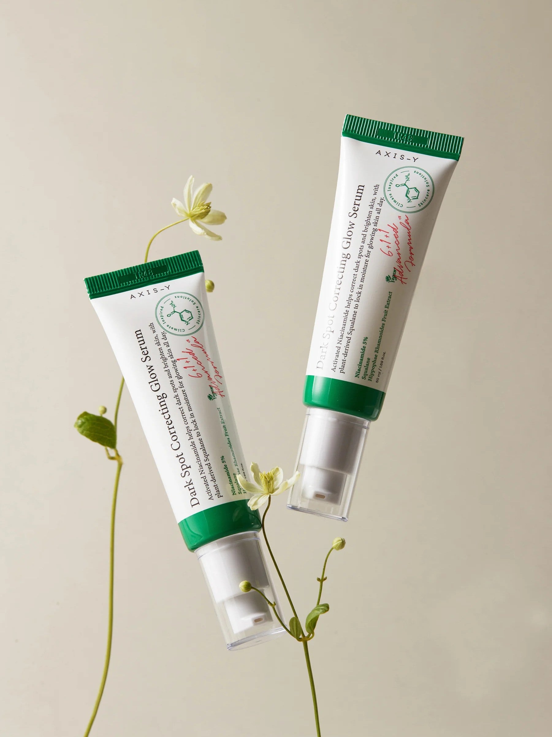 Two tubes of Axis-Y Dark Spot Correcting Glow Serum with green lids, displayed with delicate flowers on a beige background.