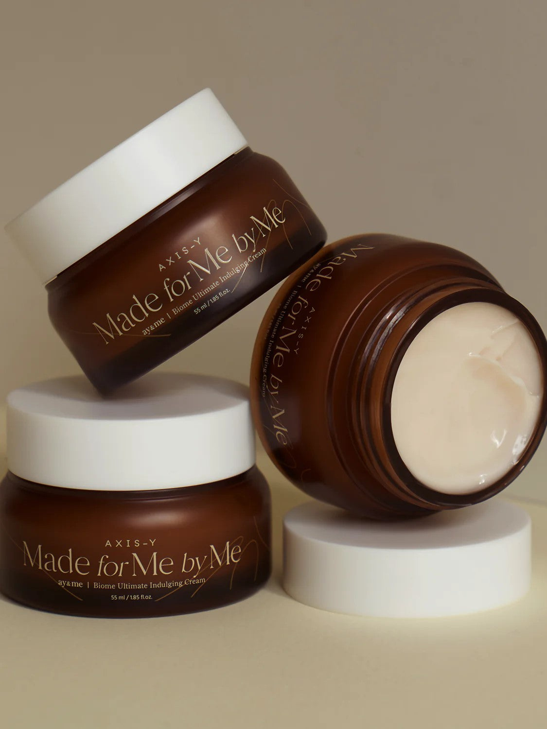 Three brown jars of AXIS-Y 'Made for Me by Me' Biome Ultimate Indulging Cream, elegantly stacked against a beige background.
