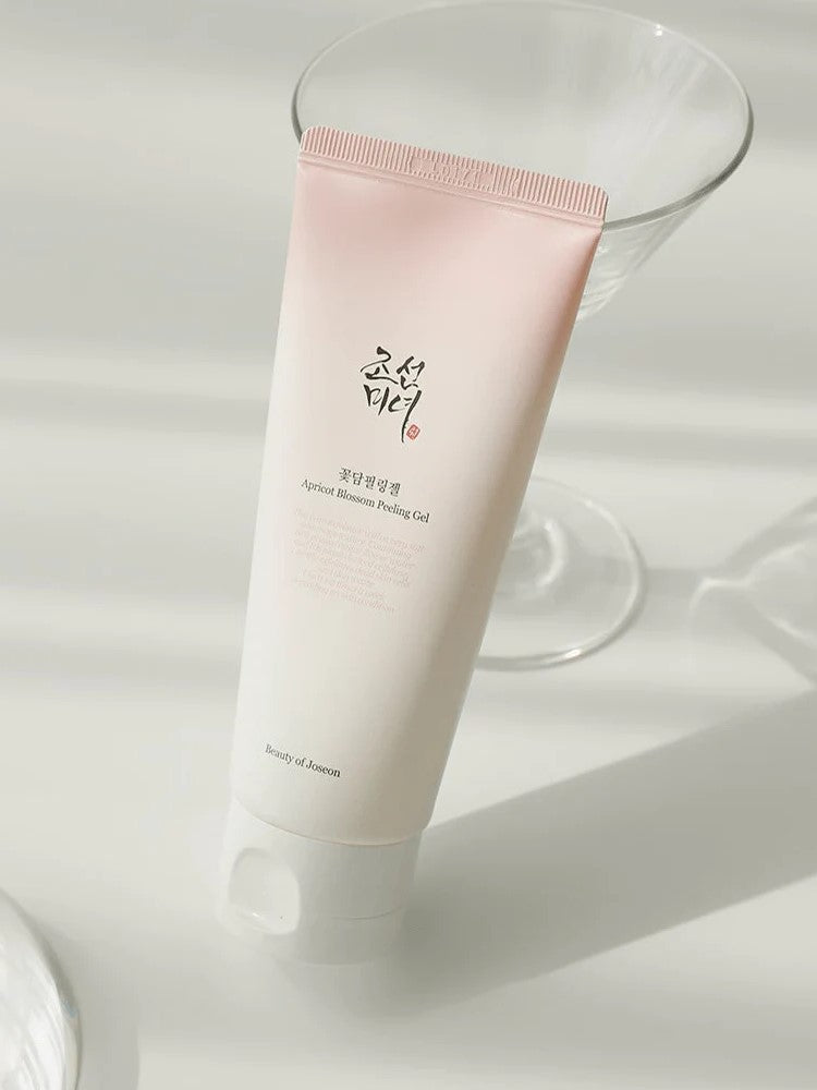 A tube of Apricot Blossom Peeling Gel from Beauty of Joseon, elegantly positioned on a reflective surface, with a glass in the background to enhance its aesthetic appeal.