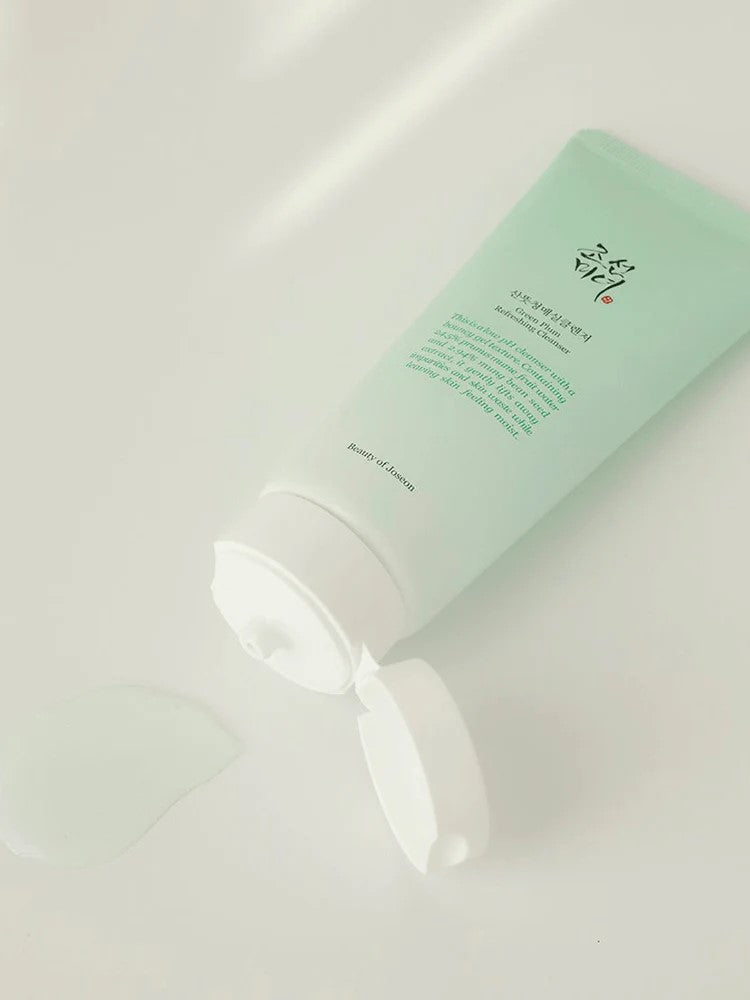 Beauty of Joseon Green Plum Refreshing Cleanser lying on a reflective surface with the cap open and a small amount of light gel texture oozing out, showcasing its refreshing formula.