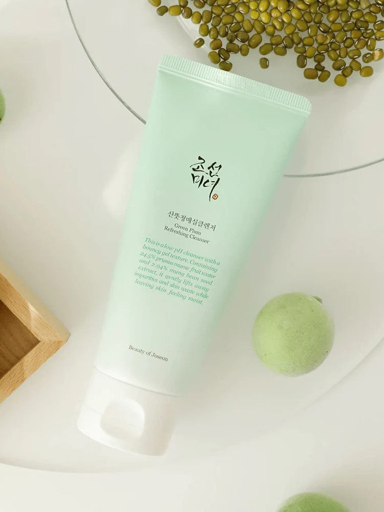 A mint-green tube of Beauty of Joseon Green Plum Refreshing Cleanser placed on a light surface, surrounded by fresh green plums and seeds, highlighting its natural ingredients.
