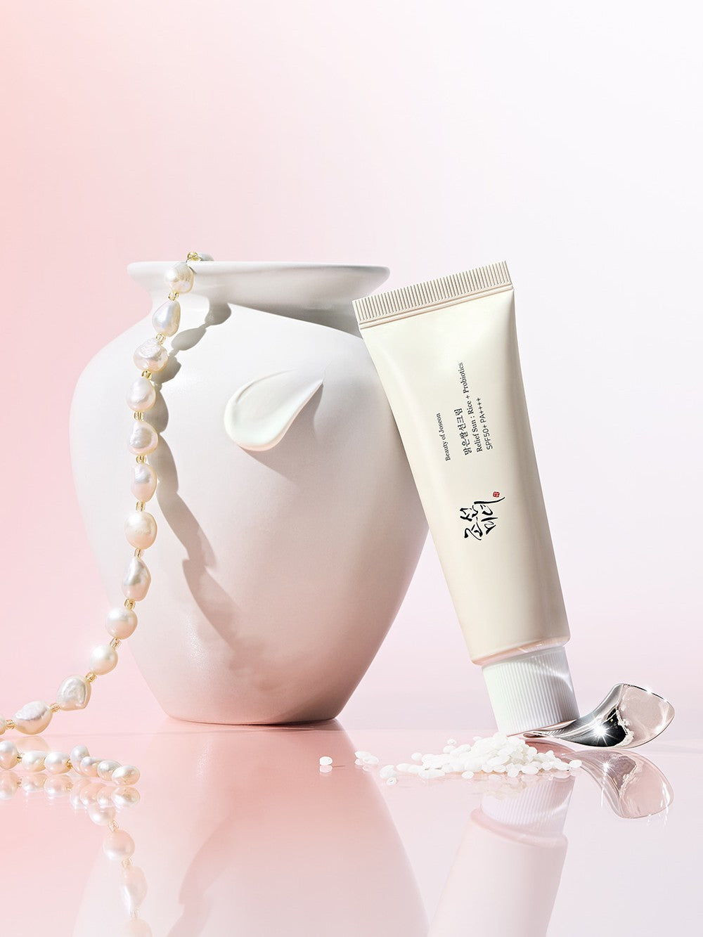 Beauty of Joseon Relief Sun: Rice + Probiotics leaning against a white vase, surrounded by pearls and cosmetic particles on a soft pink background, highlighting its elegant design and skincare benefits.