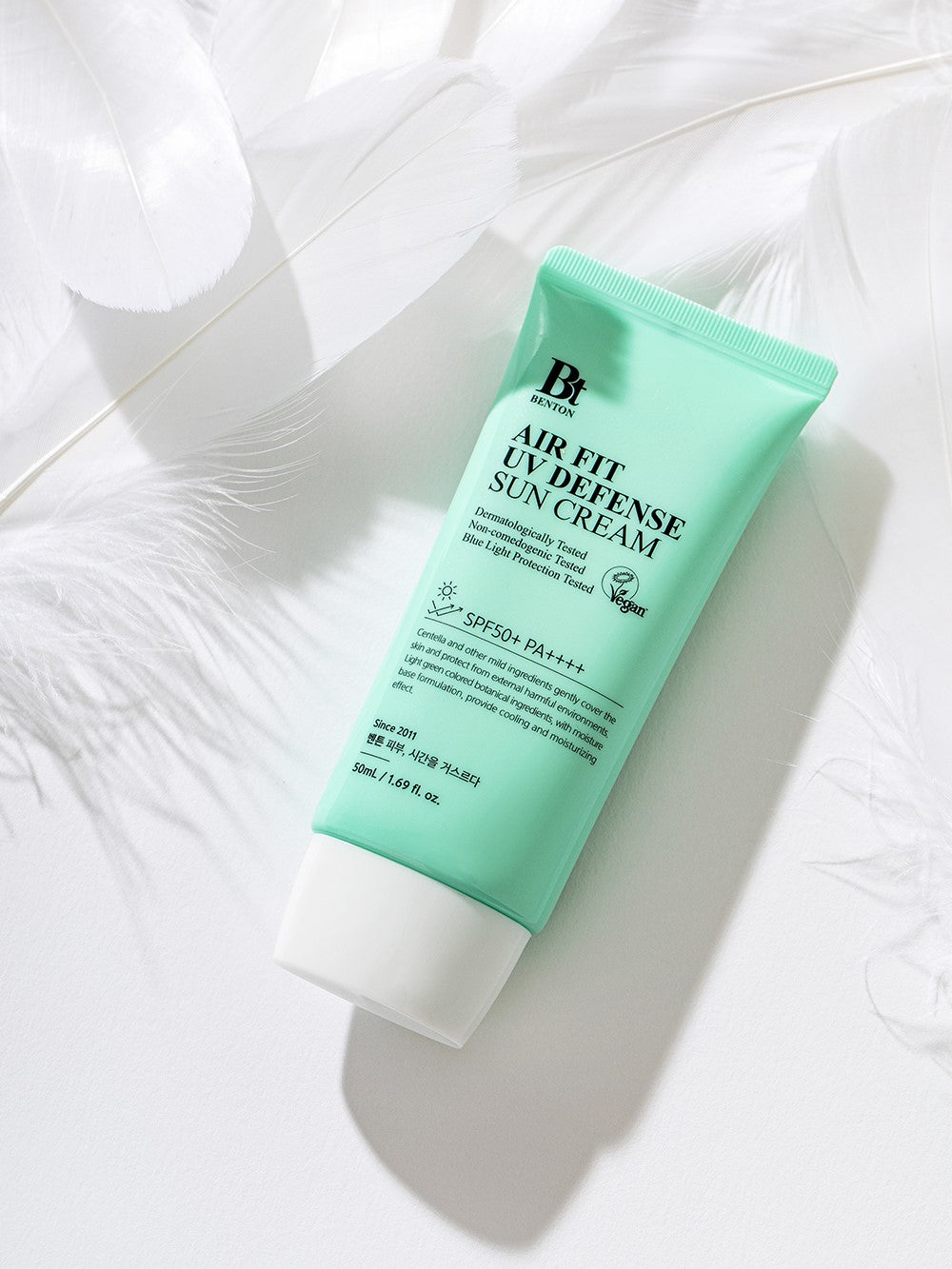A mint green tube of Benton Air Fit UV Defense Sun Cream lying on a white background with feathers.