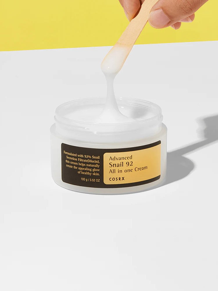 A hand holding a wooden spatula dips into a jar of Cosrx Advanced Snail 92 All in One Cream, showcasing its smooth texture against a yellow background.