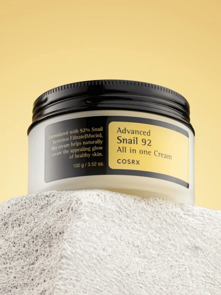 A jar of Cosrx Advanced Snail 92 All in One Cream displayed on a textured surface, emphasizing its snail mucin formula for a radiant skin glow.
