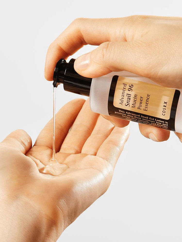 A hand holds a bottle of Cosrx Advanced Snail 96 Mucin Power Essence, dispensing its clear, gel-like texture into the palm below.