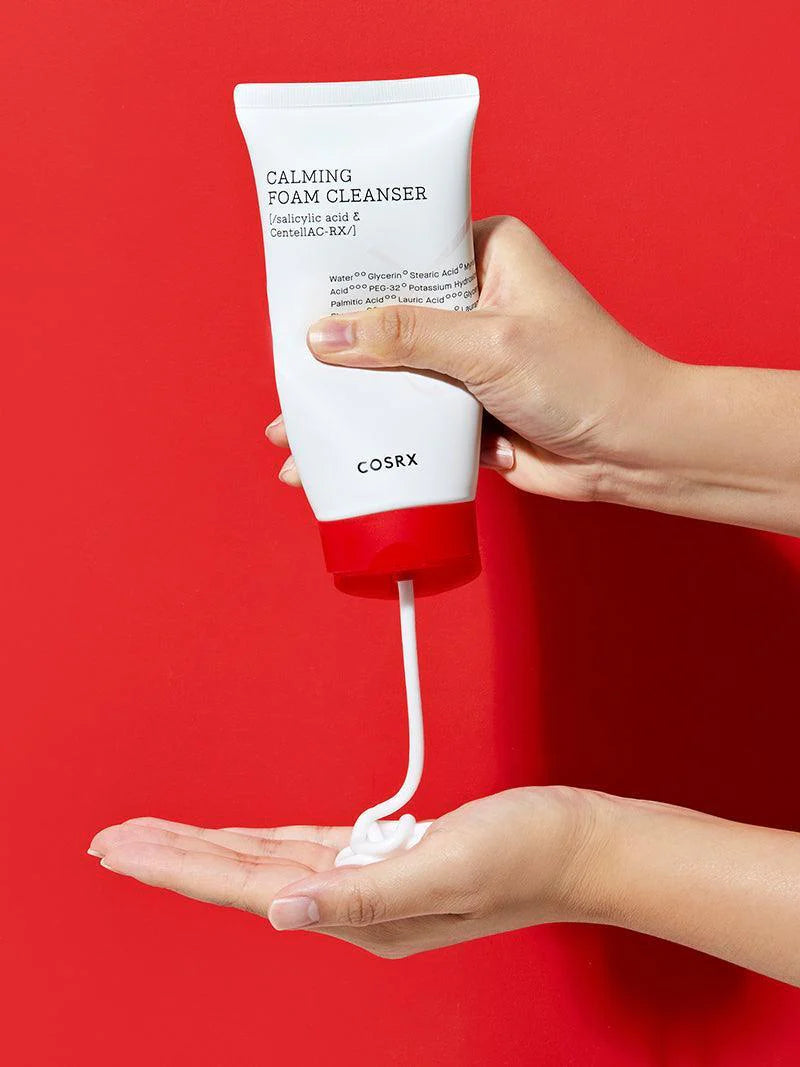 A hand dispenses creamy foam from a white tube of Cosrx Calming Foam Cleanser against a vibrant red background.
