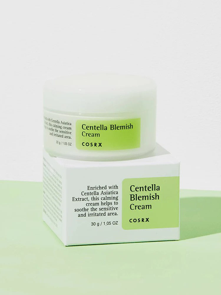 A jar of Cosrx Centella Blemish Cream with a white lid and green label, resting on its packaging against a white and green backdrop.