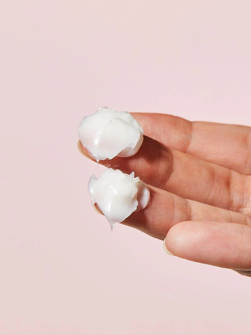 A hand holds two dollops of Cosrx Ceramide Cream, highlighting its creamy texture against a soft pink background.