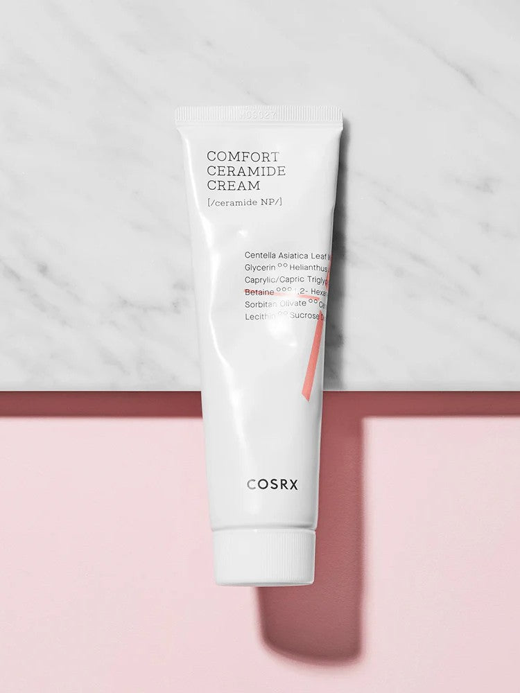 A tube of Cosrx Comfort Ceramide Cream, featuring a minimalist design, sits on a marble surface against a pink background.