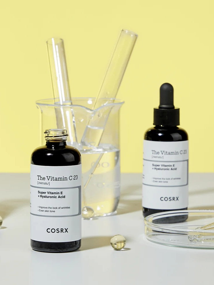 Two black bottles of Cosrx The Vitamin C 23 Serum on a yellow background, paired with glass beakers and pipettes, evoking skincare preparation.