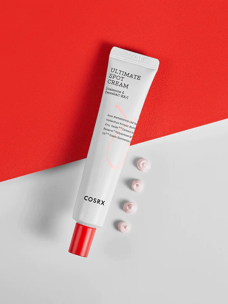 A tube of Cosrx Ultimate Spot Cream with a red cap, placed on a red and white background, alongside small dollops of cream.