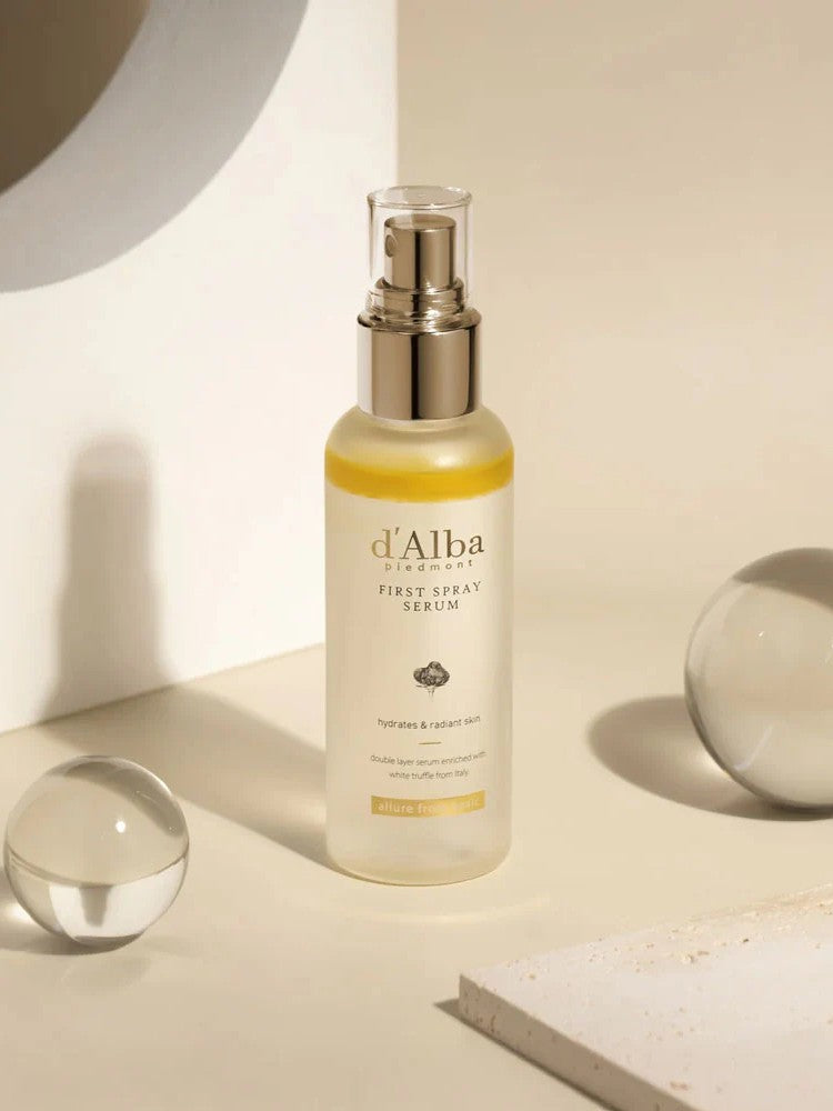 A bottle of d'Alba First Spray Serum on a minimalist surface with glass spheres, emphasizing hydration and radiance.