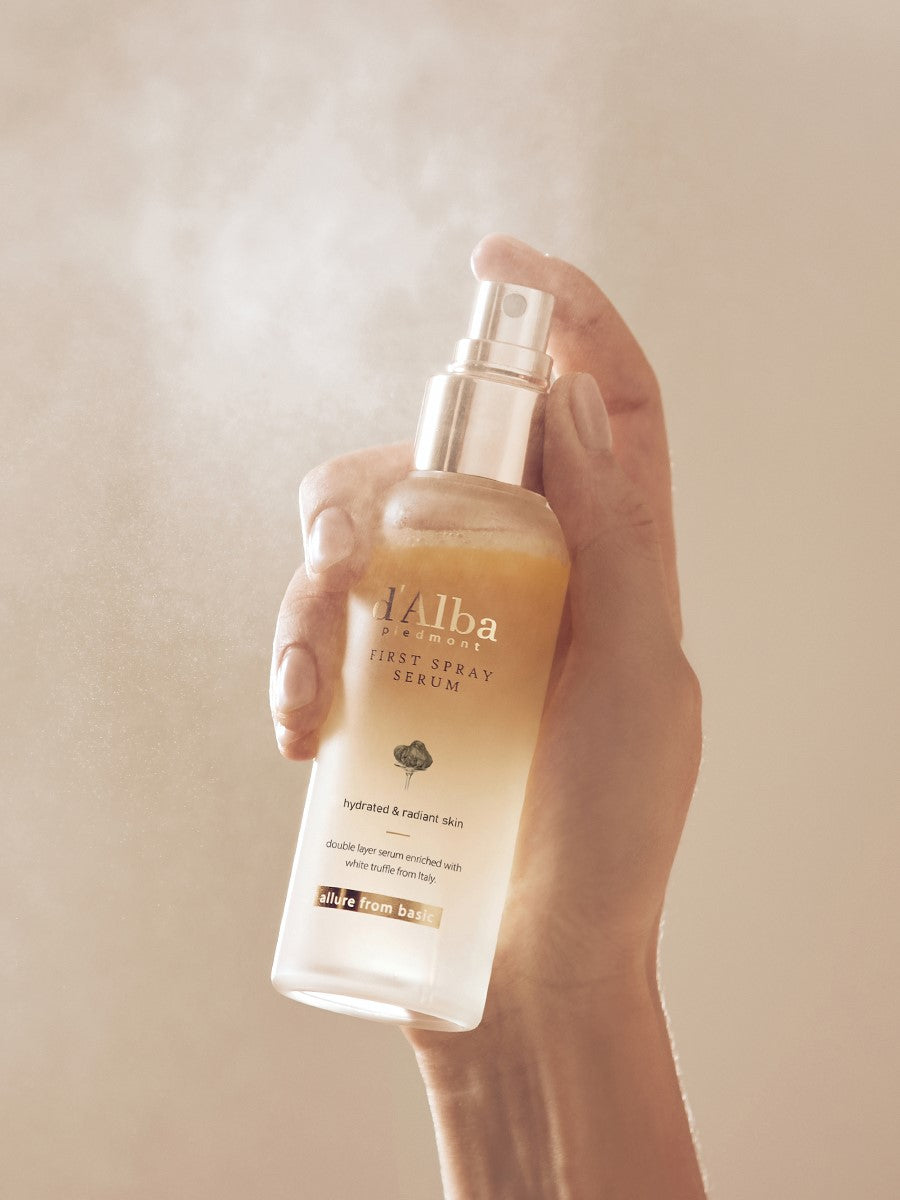 A hand holds a d'Alba First Spray Serum bottle, releasing a fine mist to highlight its hydrating and radiance-boosting benefits.