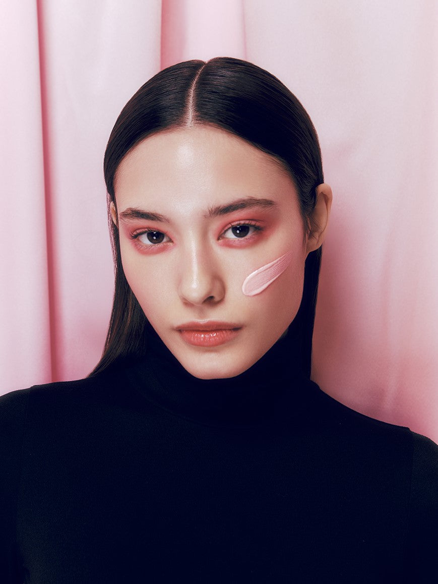 A model with sleek, straight hair wearing a black turtleneck, showcasing smooth, glowing pink cream texture of d'Alba Waterfull Tone-Up Sunscreen on her cheek, standing against a soft pink curtain background.