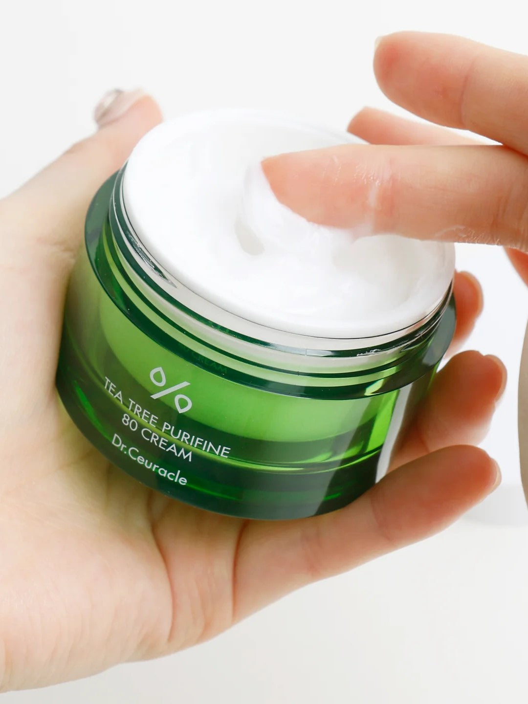 A hand dipping into a green jar of Dr. Ceuracle Tea Tree Purifine 80 Cream, showcasing its creamy texture.