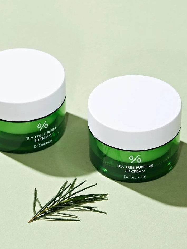 Two green jars of Dr. Ceuracle Tea Tree Purifine 80 Cream on a pale green surface, accompanied by fresh tea tree leaves, highlighting its soothing and clarifying properties.