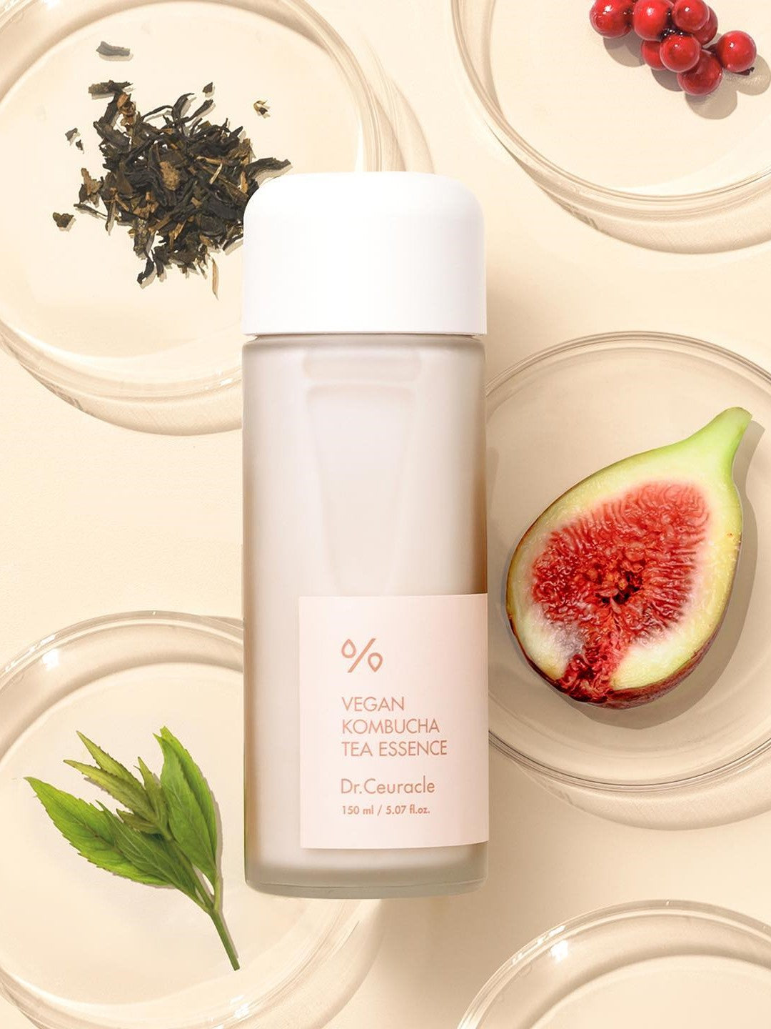 A bottle of Dr. Ceuracle Vegan Kombucha Tea Essence, surrounded by Kombucha Tea leaves, a halved fig, Schisandra chinensis berries, and assorted leaves, set against a cream background.