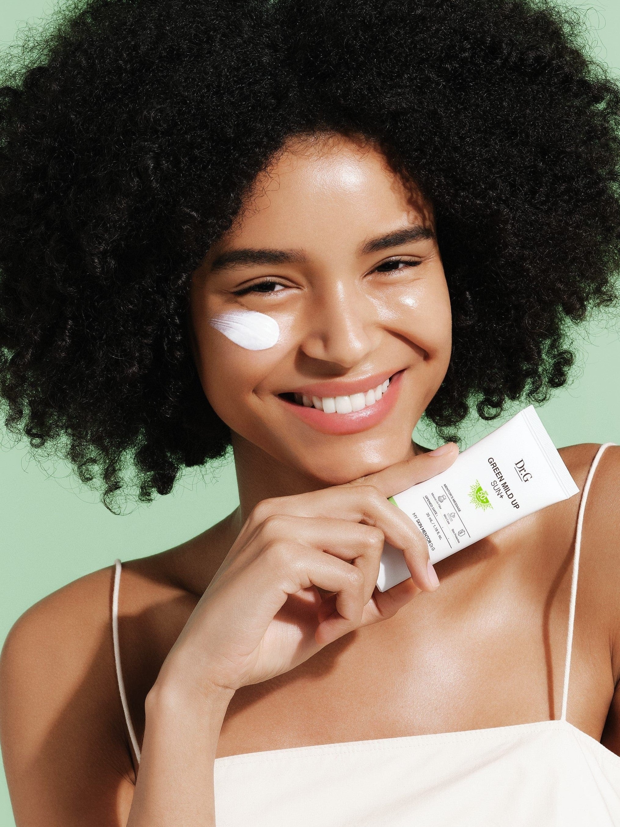 A model holding Dr.G Green Mild Up Sun+ Sunscreen, with the product applied on her cheek against a soft green background.
