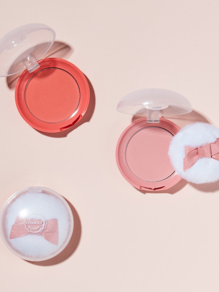 Two Etude House Lovely Cookie Blusher compacts in coral and pink, each topped with a fluffy applicator, arranged on a soft pink background.