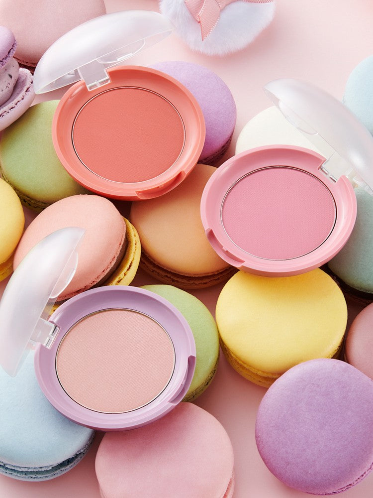 Three colorful blush compacts from the Etude House Lovely Cookie Blusher collection placed on pastel macarons, featuring peach, pink, and lavender shades against a soft pink background.