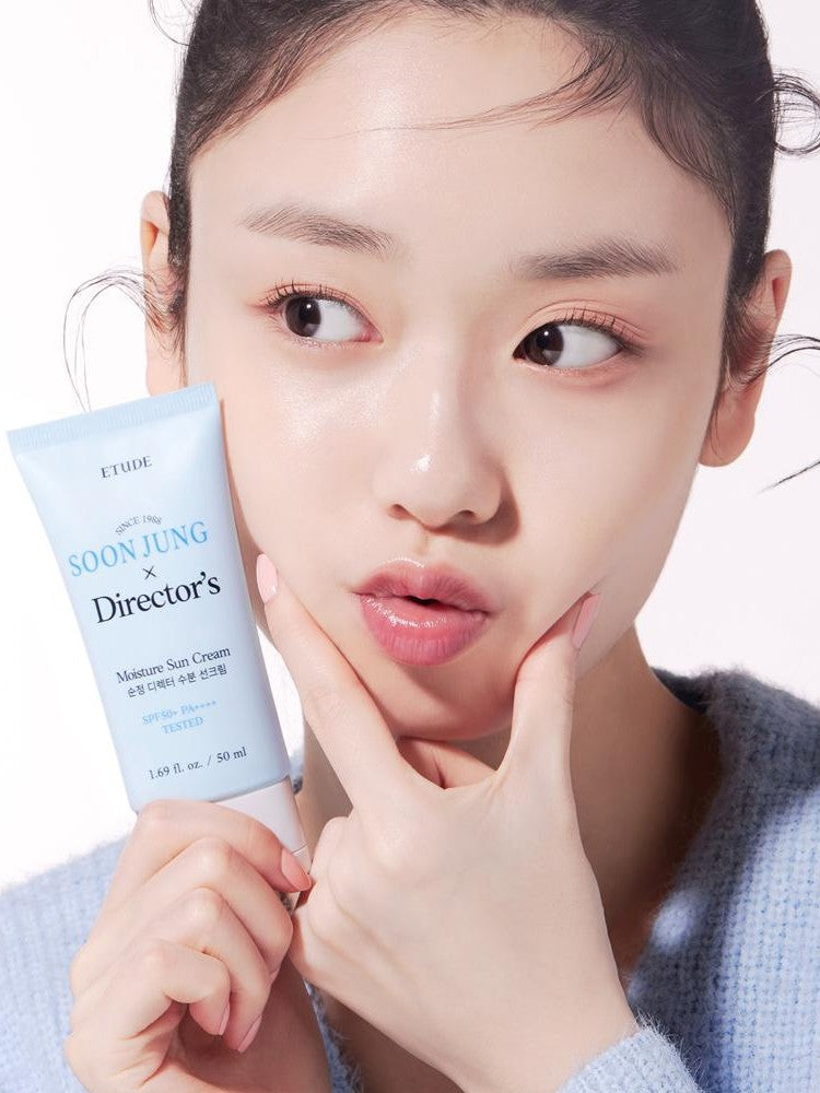 A model holding a tube of Etude House Soon Jung Director Moisture Sun Cream, wearing a light blue sweater in soft lighting.