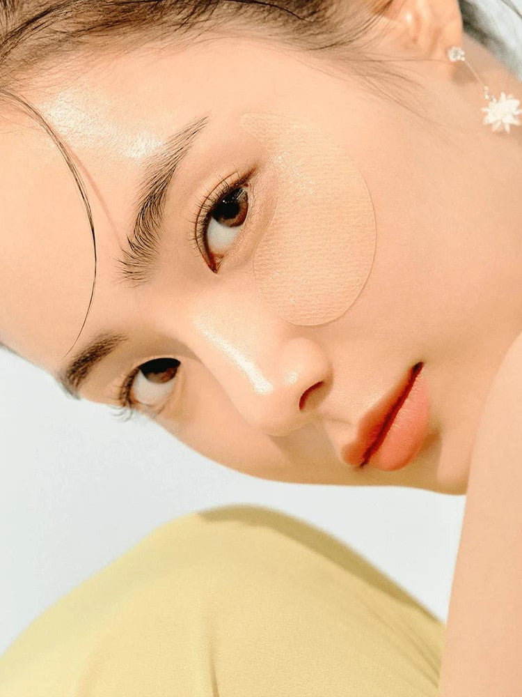 A close-up view of a model with wavy brown hair, featuring a Goodal Green Tangerine glossy yellow gel patch applied under one eye, set against a soft, blurred background.
