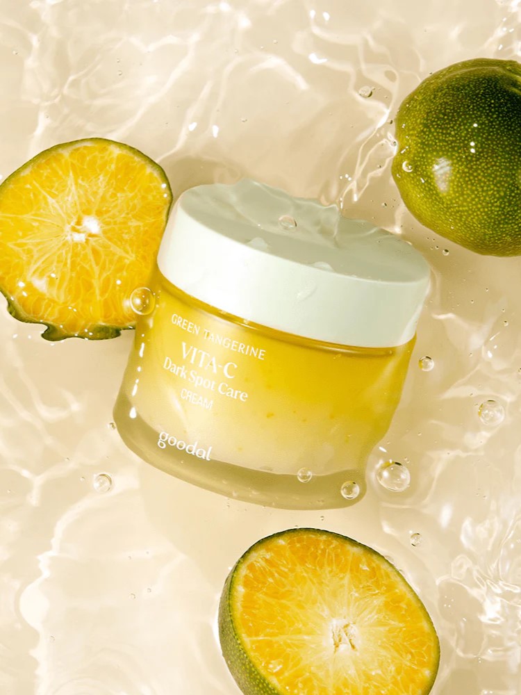A jar of Goodal Vita-C Dark Spot Care Cream surrounded by fresh green tangerines, its main ingredient, floating against a light, watery background.