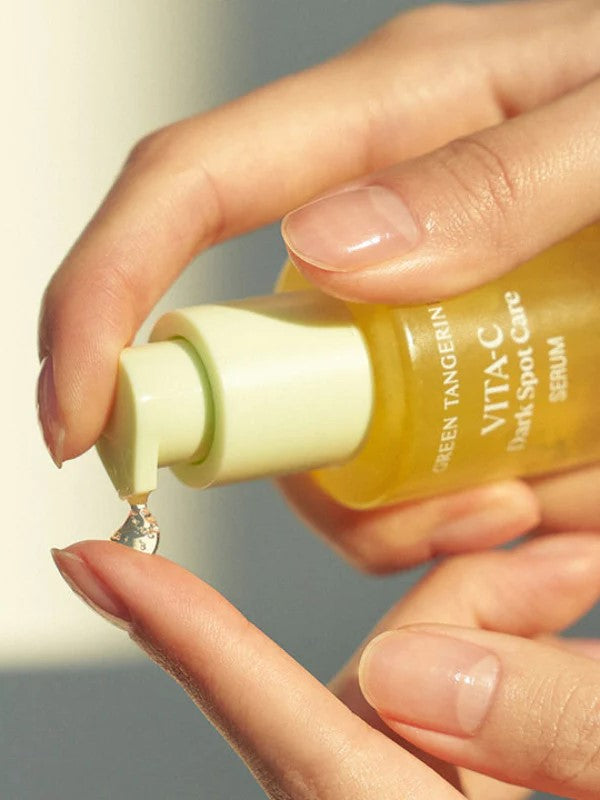A hand holding the Goodal Green Tangerine Vita-C Dark Spot Care Serum bottle, dispensing a drop onto a fingertip in soft, natural lighting.