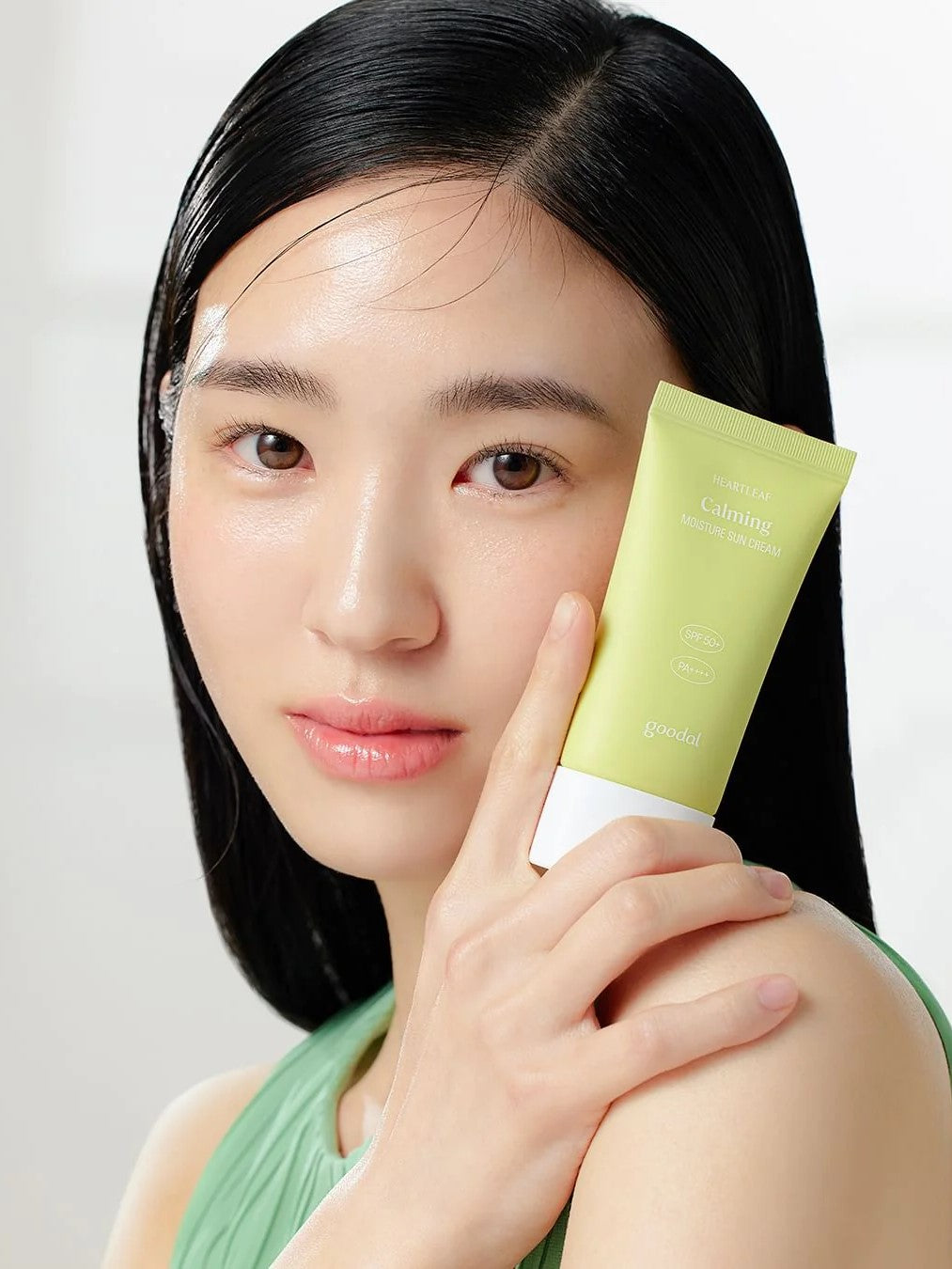 A model with sleek black hair, wearing a green top, holds a tube of Goodal Houttuynia Cordata Calming Moisture Sun Cream against her shoulder in soft lighting.