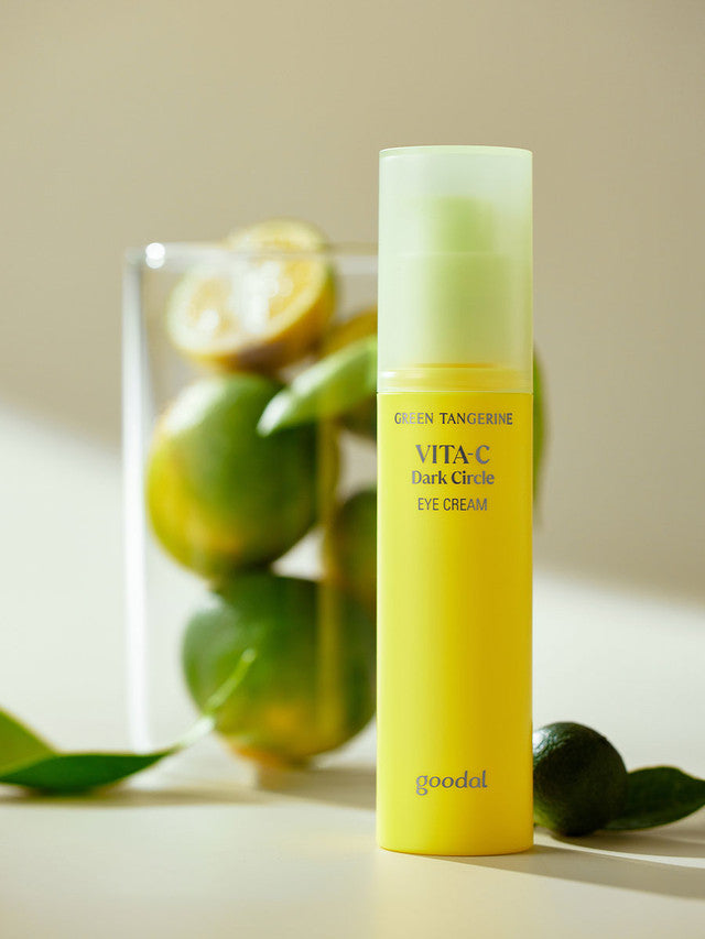 A bright yellow bottle of Goodal Green Tangerine Vita C Dark Circle Eye Cream, set on a soft, light surface with green tangerines in the background, highlighting its main ingredient.