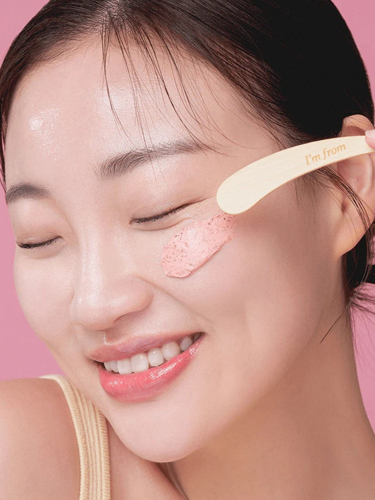 A model applying 'I'm From Beet Purifying Mask' on her cheek with its wooden spatula, showcasing its creamy and rich texture for purifying the skin.