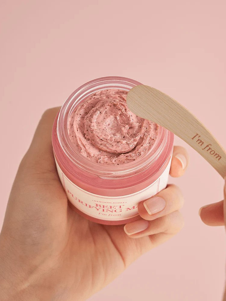 A hand holding a jar of 'I'm From Beet Purifying Mask' pink cream with a wooden spatula hovering above, set against a soft pink background, highlighting its creamy, antioxidant-rich formula.