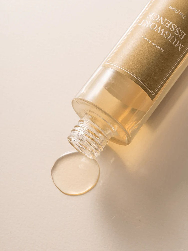 A bottle of 'I'm From' Mugwort Essence tipped over, with a droplet of golden liquid pooling on a light surface.