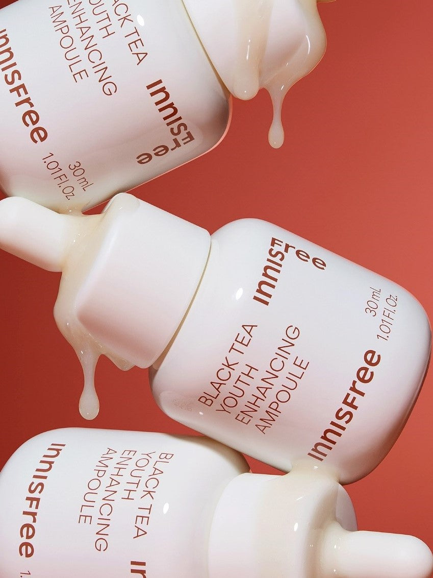 Three Innisfree Black Tea Youth Enhancing Ampoule bottles, white with amber text, dripping essence against a coral background, highlighting its luxurious and rejuvenating formula.