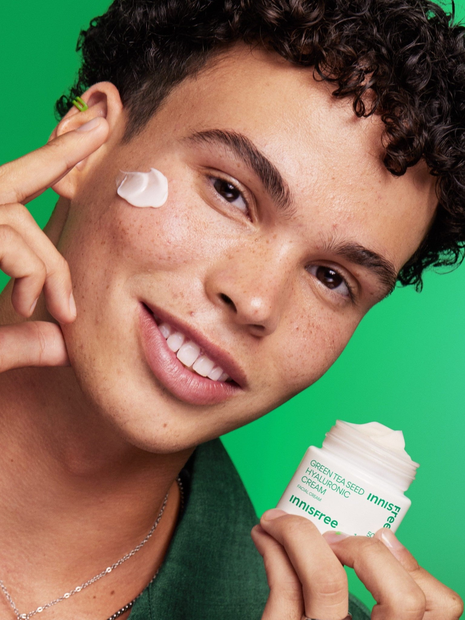 A male model holding a jar of Innisfree Green Tea Seed Hyaluronic Cream against a vibrant green background, showcasing its smooth texture applied to his cheek.