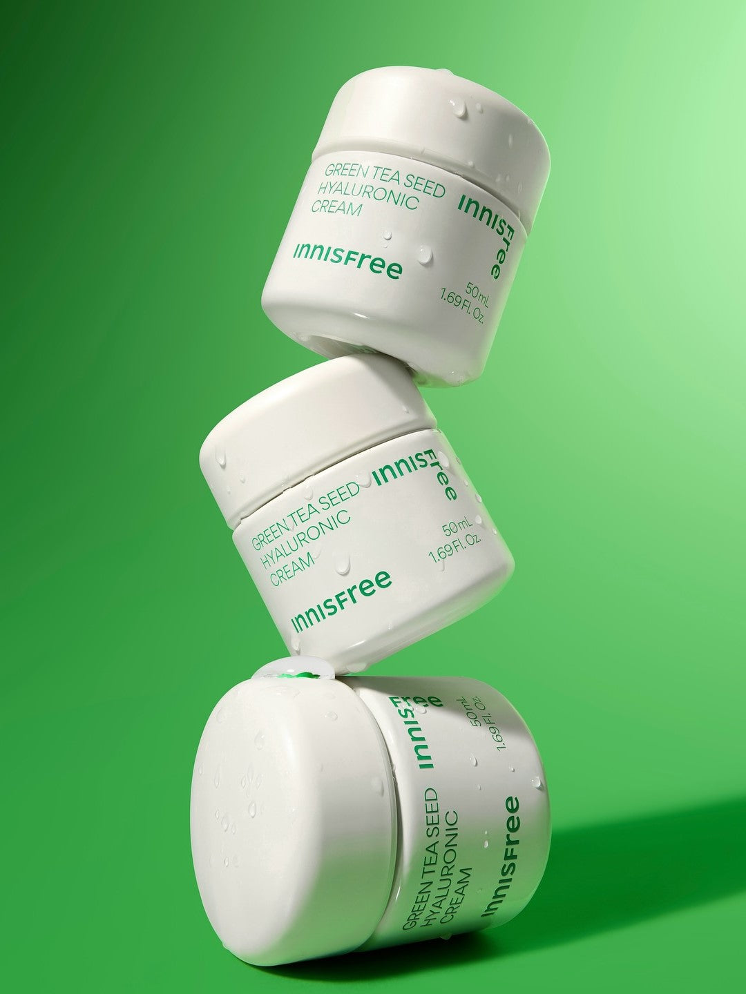 Three Innisfree Green Tea Seed Hyaluronic Cream jars stacked on a vibrant green background, glistening with droplets of water to emphasize hydration and freshness.