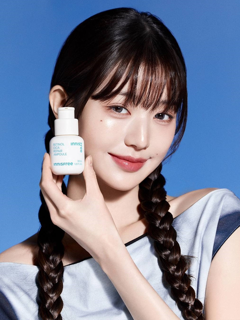 A model with long braided hair holds an Innisfree Retinol Cica Repair Ampoule against a bright blue background.