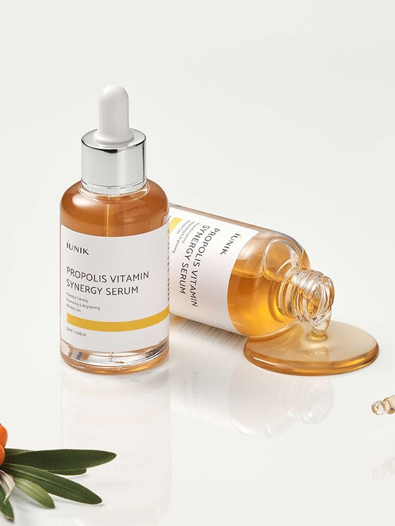 Bottle of iUNIK Propolis Vitamin Synergy Serum with golden liquid spilling from dropper, surrounded by green leaves and sea buckthorn fruit, highlighting nourishing and brightening benefits.